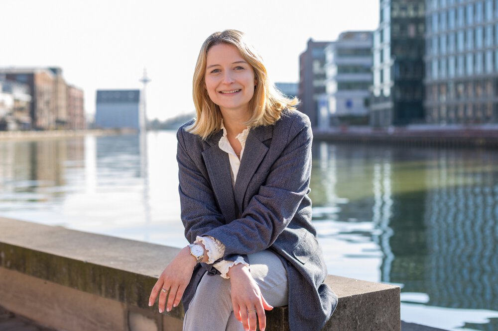 Eine Frau mit blonden Haaren und einem grauen Jackett sitzt lächelnd auf einer Mauer vor einem Hafengelände mit zahlreichen Neubauten.