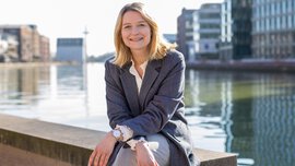 Eine Frau mit blonden Haaren und einem grauen Jackett sitzt lächelnd auf einer Mauer vor einem Hafengelände mit zahlreichen Neubauten.