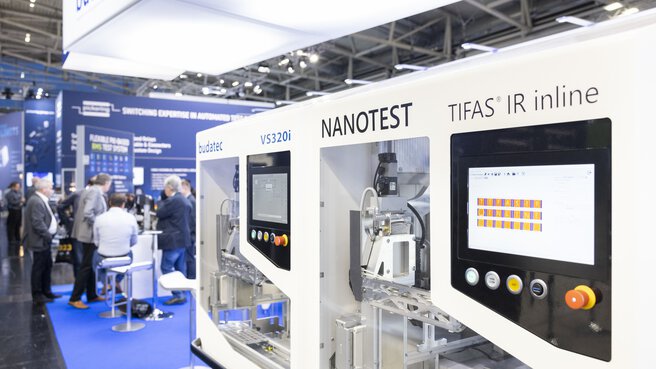 Ausgestellte Maschinen an einem Stand auf der productronica. Im Hintergrund steht eine Gruppe Menschen beisammen.