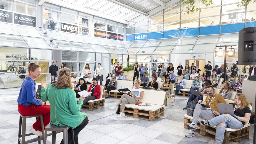 People sit on comfortable benches made of wooden pallets in a modern interior and listen to a lecture by two women on bar stools.