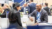 A black-haired young woman and a dark-skinned man are talking at the Transport and Logistic 2025 trade fair in Munich. In the background, many people can be seen networking.