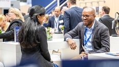 Auf der Messe Transport und Logistic 2025 in München unterhalten sich eine schwarzhaarige junge Frau und ein dunkelhäutiger Mann. Im Hintergrund sieht man viele Menschen umschafft beim Netzwerken.