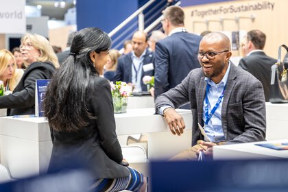 Auf der Messe Transport und Logistic 2025 in München unterhalten sich eine schwarzhaarige junge Frau und ein dunkelhäutiger Mann. Im Hintergrund sieht man viele Menschen umschafft beim Netzwerken.