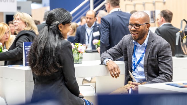 A black-haired young woman and a dark-skinned man are talking at the Transport and Logistic 2025 trade fair in Munich. In the background, many people can be seen networking.