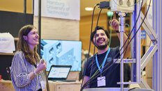 Ein Universitätsmitarbeiter erklärt einer Messebesucherin mit Begeisterung eine technische Versuchsanlage.