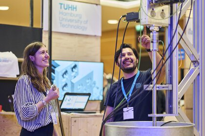 Ein Universitätsmitarbeiter erklärt einer Messebesucherin mit Begeisterung eine technische Versuchsanlage.