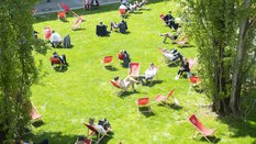 Besucher der transport logistic entspannen sich in zahlreichen roten Liegestühlen auf einer sonnenbeschienenen Grünfläche zwischen den Messehallen.