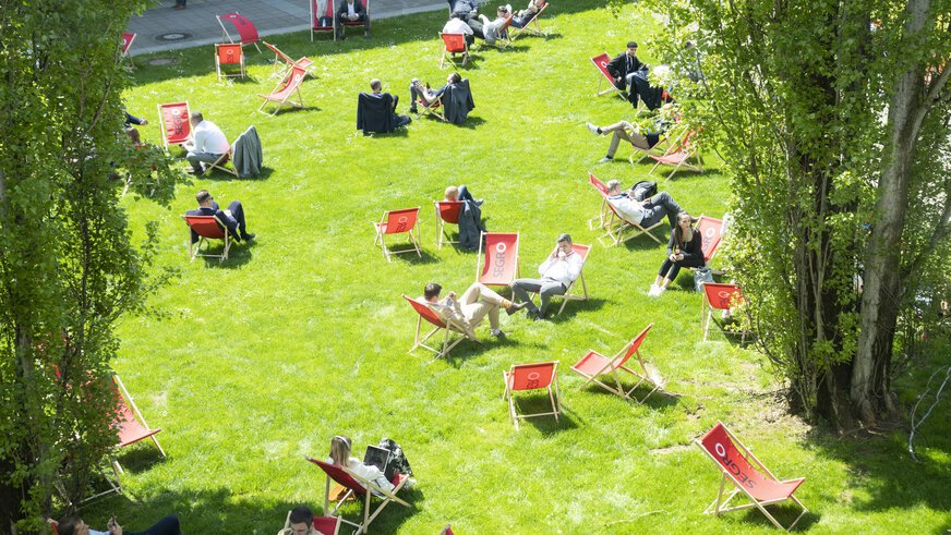 Besucher der transport logistic entspannen sich in zahlreichen roten Liegestühlen auf einer sonnenbeschienenen Grünfläche zwischen den Messehallen.