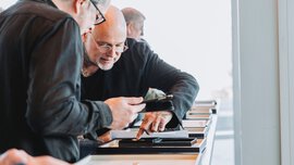 Two men dressed in black and wearing glasses are looking closely at a piece of jewelry in a black wooden box.