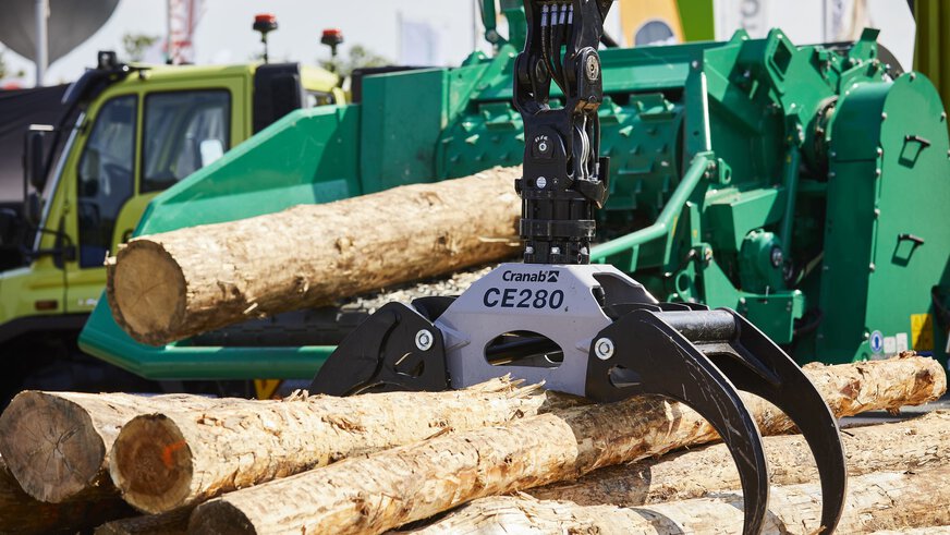 The gripper arm of a forestry machine with black claws surrounds several tree trunks, in the background a tree trunk lies on a green wood chipper.