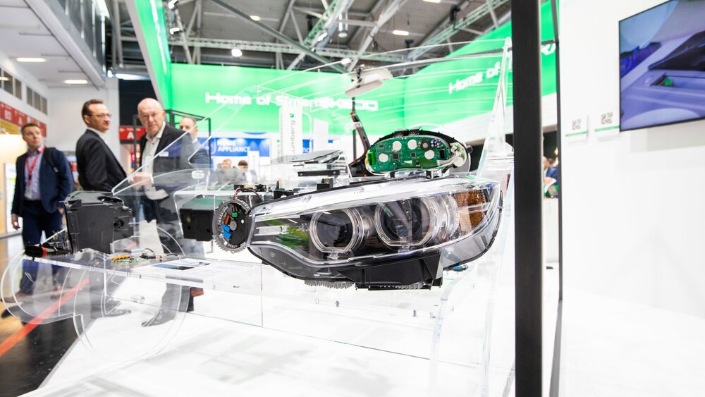 A vehicle headlight is mounted in a transparent model of a car at a trade fair stand. Visitors can be seen in the background.