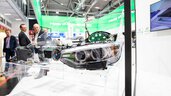 Ein Fahrzeugscheinwerfer ist an einem Messestand in einem transparenten Modell eines Autos montiert. Im Hintergrund sind Besucher zu sehen.