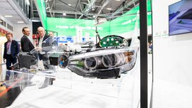 A vehicle headlight is mounted in a transparent model of a car at a trade fair stand. Visitors can be seen in the background.