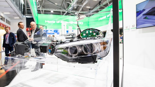 Ein Fahrzeugscheinwerfer ist an einem Messestand in einem transparenten Modell eines Autos montiert. Im Hintergrund sind Besucher zu sehen.