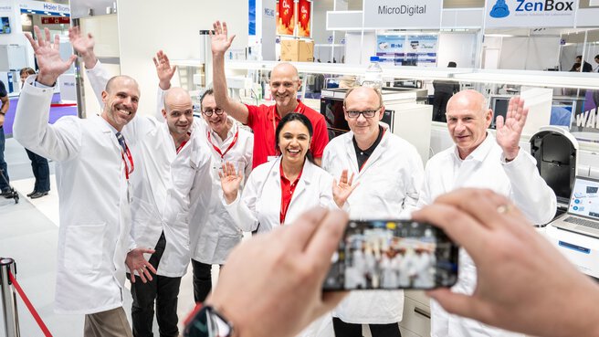 A cell phone takes a photo of seven smiling people in lab coats.