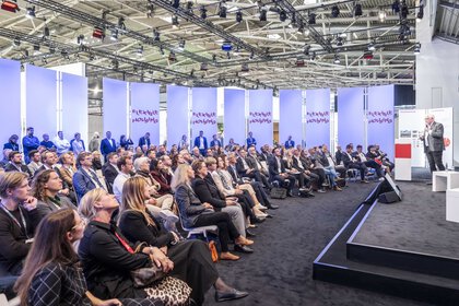 Ein großes Publikum sitzt in mehreren Reihen vor einer Bühne auf der Expo Real und hört einem Sprecher zu, der rechts im Bild am Podium steht. Hinter dem Publikum stehen weitere Besucher. Im Hintergrund sind hohe, beleuchtete Paneele mit der Aufschrift „Flexible Housing“ zu sehen. Die Halle ist hell ausgeleuchtet und zeigt Messecharakter.