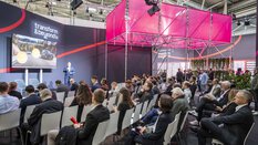A presentation on the Tech Talk stage at EXPO REAL in Munich. A speaker stands in front of a large screen displaying a presentation about the electric vehicle market. The audience is seated in rows, listening attentively, with some taking notes or using their smartphones. The stage features a modern design with bright pink structures and greenery in the background.