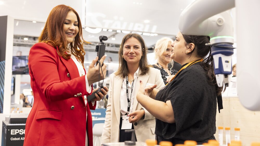 Drei Frauen unterhalten sich lebhaft an einem Messestand, eine von ihnen hält ein Kamera-Gimbal in der Hand. Im Vordergrund sind Laborfläschchen und ein Roboterarm zu sehen. Die Szene vermittelt eine freundliche und technologieorientierte Atmosphäre.