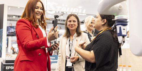Three women are engaged in a lively conversation at a trade fair booth, with one holding a camera gimbal. In the foreground, lab vials and a robotic arm are visible. The scene conveys a friendly, tech-focused atmosphere.