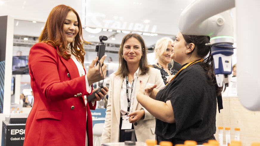Drei Frauen unterhalten sich lebhaft an einem Messestand, eine von ihnen hält ein Kamera-Gimbal in der Hand. Im Vordergrund sind Laborfläschchen und ein Roboterarm zu sehen. Die Szene vermittelt eine freundliche und technologieorientierte Atmosphäre.