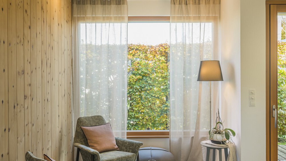 Gemütliches Zimmer mit einem Stuhl, einer Lampe und einem Beistelltisch neben einem Fenster mit durchsichtigen Vorhängen und grünem Laub draußen.