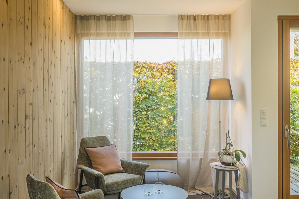 Gemütliches Zimmer mit einem Stuhl, einer Lampe und einem Beistelltisch neben einem Fenster mit durchsichtigen Vorhängen und grünem Laub draußen.