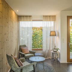 Gemütliches Zimmer mit einem Stuhl, einer Lampe und einem Beistelltisch neben einem Fenster mit durchsichtigen Vorhängen und grünem Laub draußen.