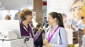 Two women at a trade fair stand with analytcia lanyards are talking in front of an open laptop.