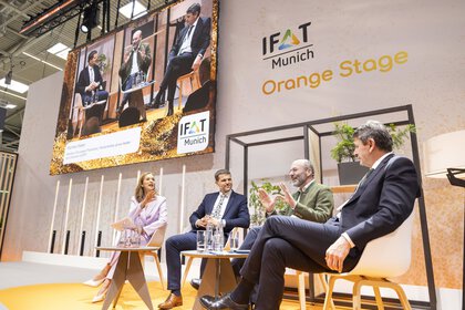 Panel discussion with four people on the stage of the IFAT Munich Orange Stage. The speakers are also shown on a large display.