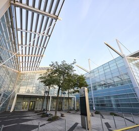 Modernes Bürogebäude aus Glas mit Metallträgern, Bäumen und klarem Himmel inmitten eines städtischen Innenhofs.
