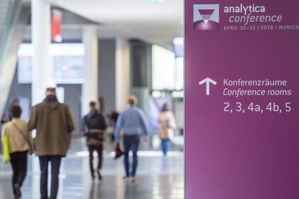 People walking in a corridor to the analytica conference, signpost in purple points to conference rooms 2, 3, 4a, 4b, 5.