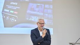 A man with white hair and glasses speaks into a microphone in front of a projection screen displaying digital images.