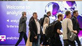 A group of people walk past a large sign in shades of purple that reads 'Welcome to analytica 2024'.