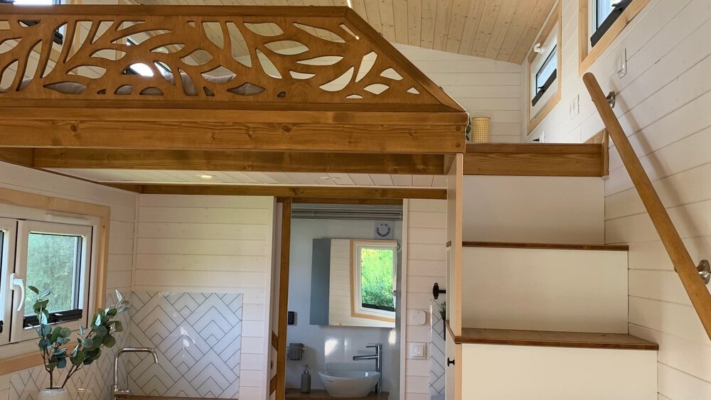 Modernes Tiny-House Interieur mit weißer Treppe, Holzgeländer und dekorativer Holzverkleidung über einem offenen Raum.