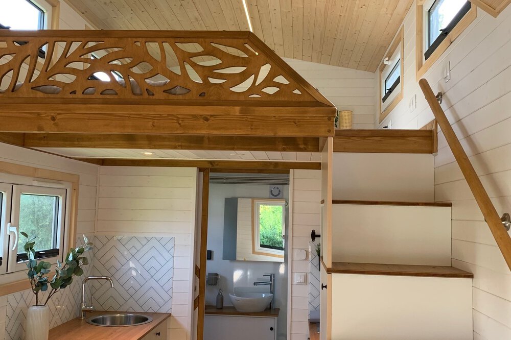 Modernes Tiny-House Interieur mit weißer Treppe, Holzgeländer und dekorativer Holzverkleidung über einem offenen Raum.