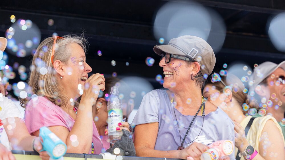 Two women smiling and blowing soap bubbles at an outdoor event, surrounded by floating soap bubbles and other people.