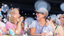 Two women smiling and blowing soap bubbles at an outdoor event, surrounded by floating soap bubbles and other people.