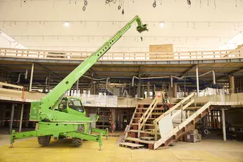 A green telescopic handler with an extended arm is on a construction site inside a hall. In the background, a multi-level wooden structure with pipes. A construction worker in safety gear stands on a staircase.