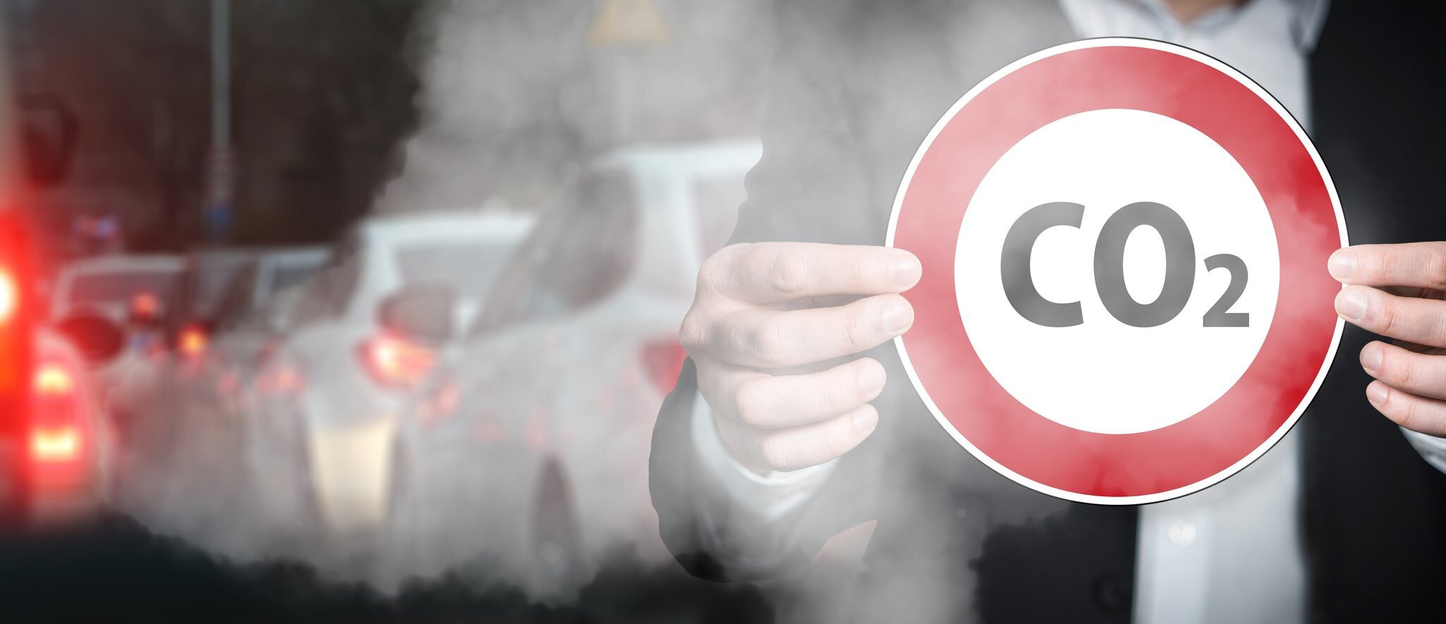 Person in a suit holds a round sign with the inscription “CO₂” in front of a smoky city scene with cars and exhaust fumes in the background.