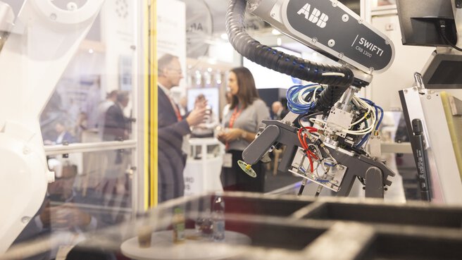 View through a glass display case in which a modern robotic arm is operating. Trade fair stands and visitors can be seen through the glass in the background.