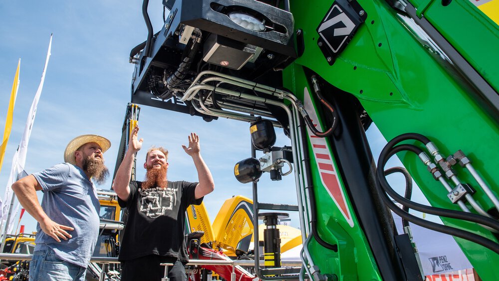 Zwei Männer stehen an einer großen grünen Maschine auf einer Outdoor-Ausstellung unter blauem Himmel.