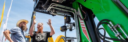 Zwei Männer stehen an einer großen grünen Maschine auf einer Outdoor-Ausstellung unter blauem Himmel.