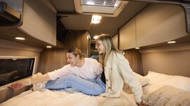 Zwei Frauen sitzen lachend auf einem Bett in einem ausgebauten Camper-Van und schauen sich begeistert um.