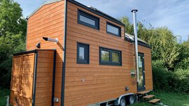 Ein kleines, hölzernes Minihaus mit mehreren Fenstern, inmitten einer grünen, belaubten Außenumgebung.