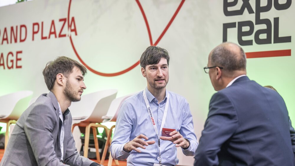Three men in suits, wearing lanyards round their necks, are sitting and chatting next to the 