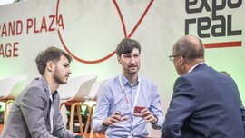 Three men in suits, wearing lanyards round their necks, are sitting and chatting next to the 