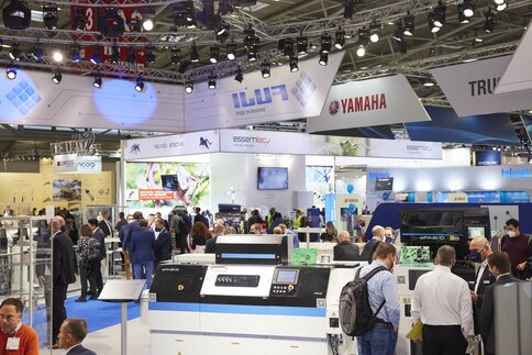 View through an event hall at productronica of various colourful exhibition stands with advertising banners and numerous interested visitors.