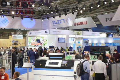 Blick durch eine Veranstaltungshalle der productronica auf diverse bunte Messestände mit Werbebannern und zahlreiche interessierte Besucher.