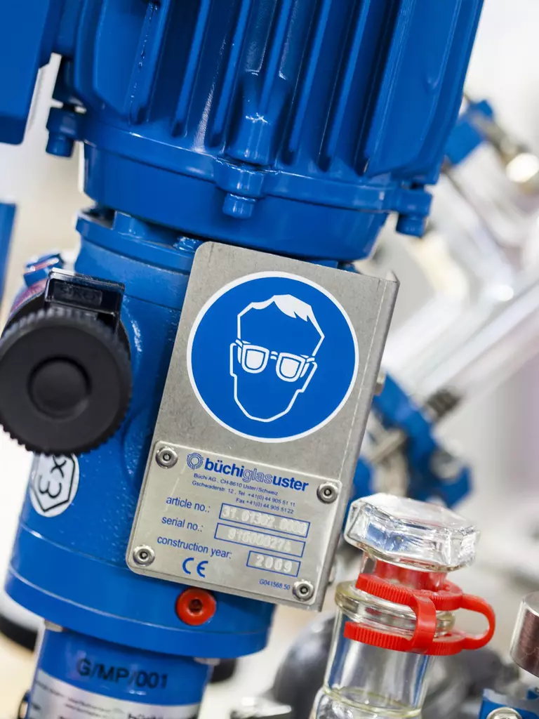 Eine Laborausrüstung mit einer blauen Vorrichtung, einem Druckmesser und verschiedenen Ventilen. Ein Warnschild für Augenschutz ist sichtbar.