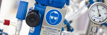 Eine Laborausrüstung mit einer blauen Vorrichtung, einem Druckmesser und verschiedenen Ventilen. Ein Warnschild für Augenschutz ist sichtbar.